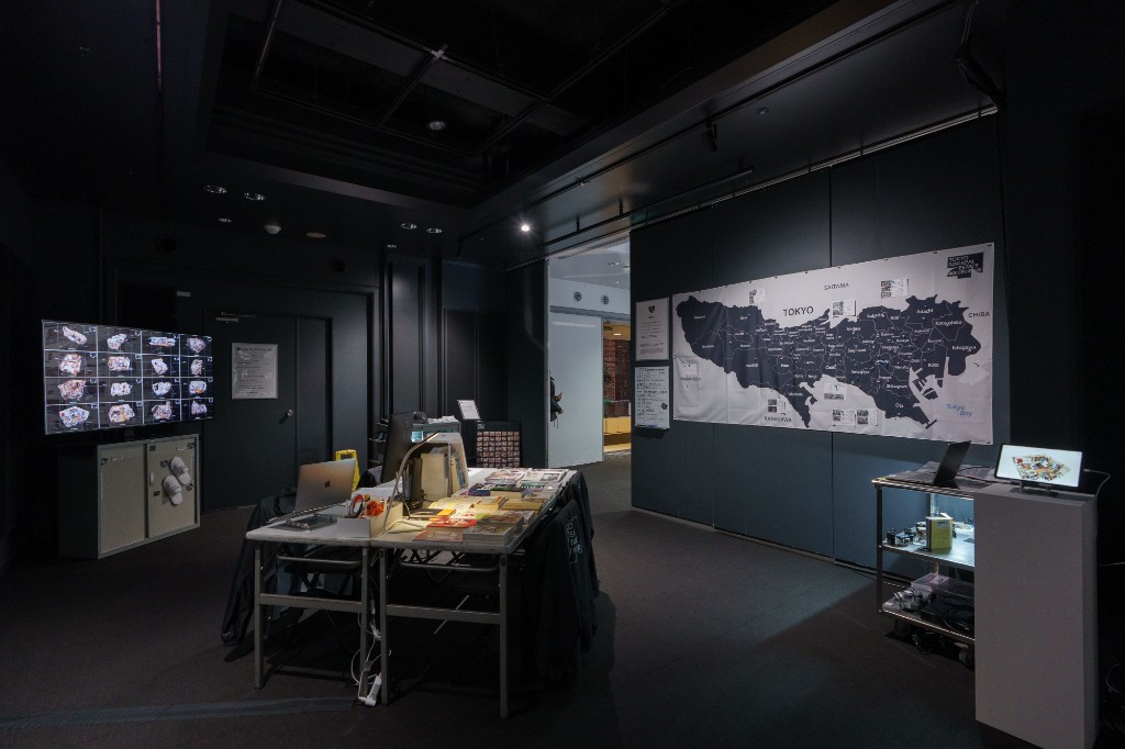 The TOKYO UNREAL ESTATE exhibition interior at CCBT showing a large Tokyo map, workstation with materials, and 3D scan displays